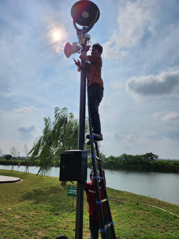 Teknisi di atas tangga memasang speaker outdoor dan kamera pada tiang di tepi danau