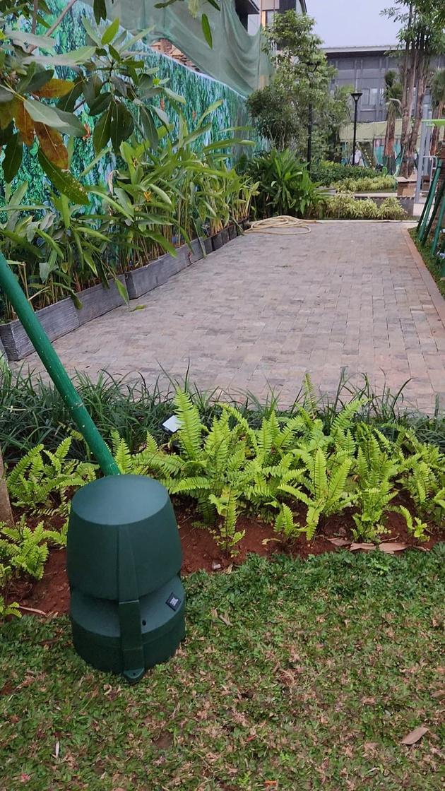 Garden speaker hijau berdiri di antara tanaman di tepi jalur pedestrian